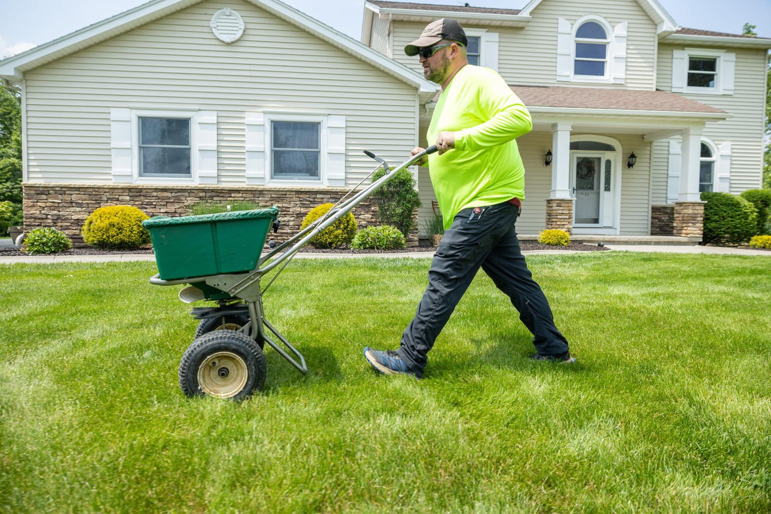 Crew member in a high-visibility shirt pushing a broadcast spreader across a front lawn